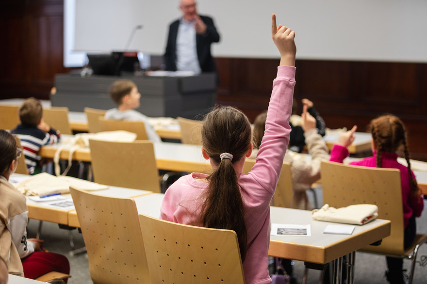 Mehrere Kinder sitzen in einem Seminarraum der Universität. Ein Mädchen meldet sich. 