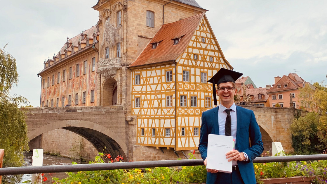 Christoph Czychon steht mit seiner Doktorarbeit vor dem Alten Rathaus in Bamberg.