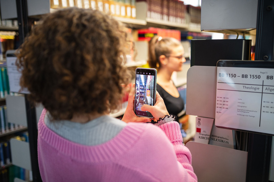 Eine Frau dreht mit dem Smartphone ein Video von einer Studentin in der Bibliothek.