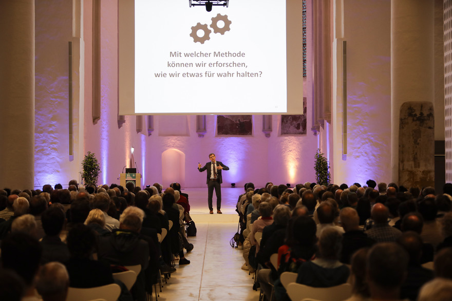 Fritz Breithaupt steht auf der Bühne. Hinter ihm eine Leinwand mit einer PowerPoint-Präsentation.