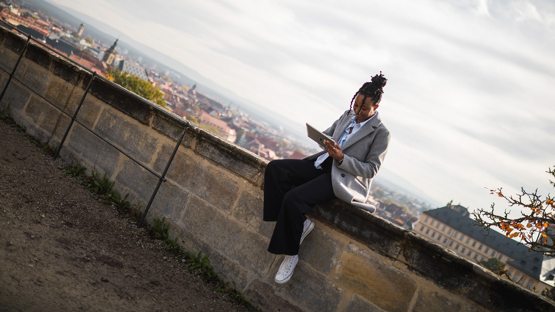 Junge Frau sitzt auf einer Mauer mit Blick auf Bamberg und bedient ein Tablet