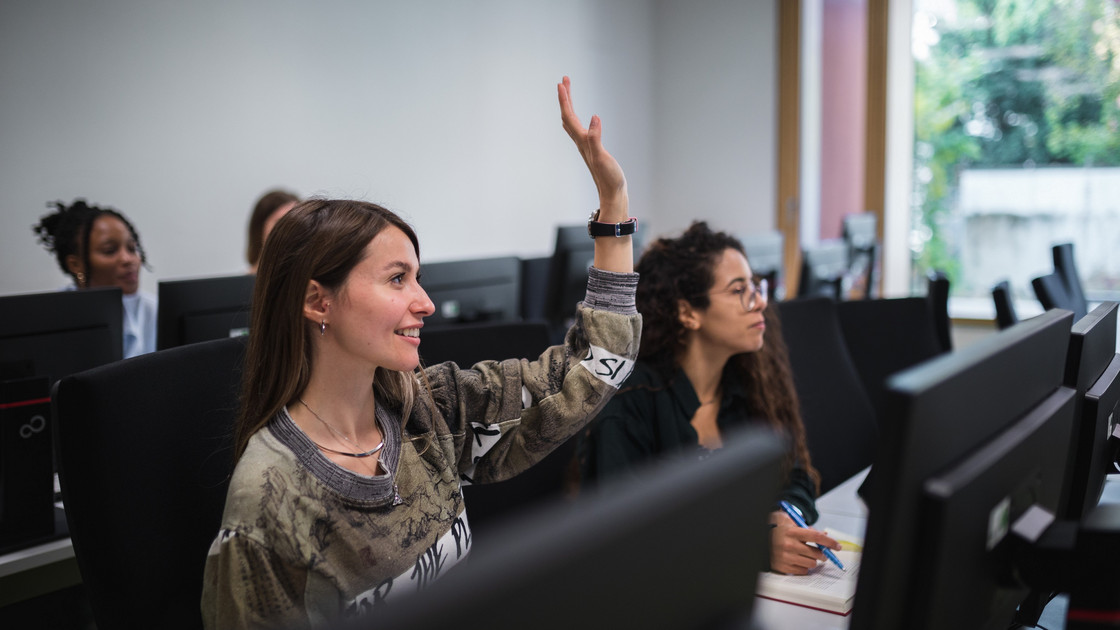 Eine junge Frau in einem Seminarraum mit Computern hebt die Hand.